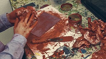 Bild: Hände voll roter Farbe und ein Spachtel auf dem Tisch