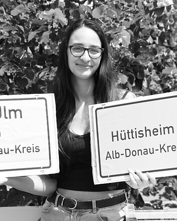 Bild: Frau hält Ortsschild Hüttisheim  und Schild vh Ulm im Alb-Donau-Kreis